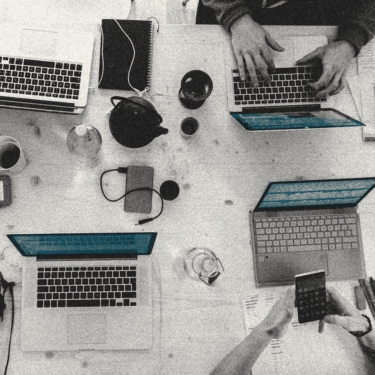 Une table de travail partagée, vue du dessus : plusieurs ordinateurs ouverts, des mains au clavier et au téléphone.